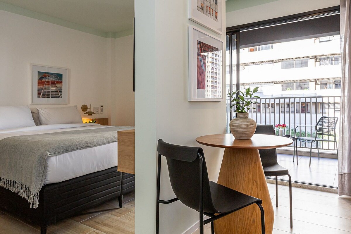 A modern hotel room with a bed, a desk, and a balcony with a view.