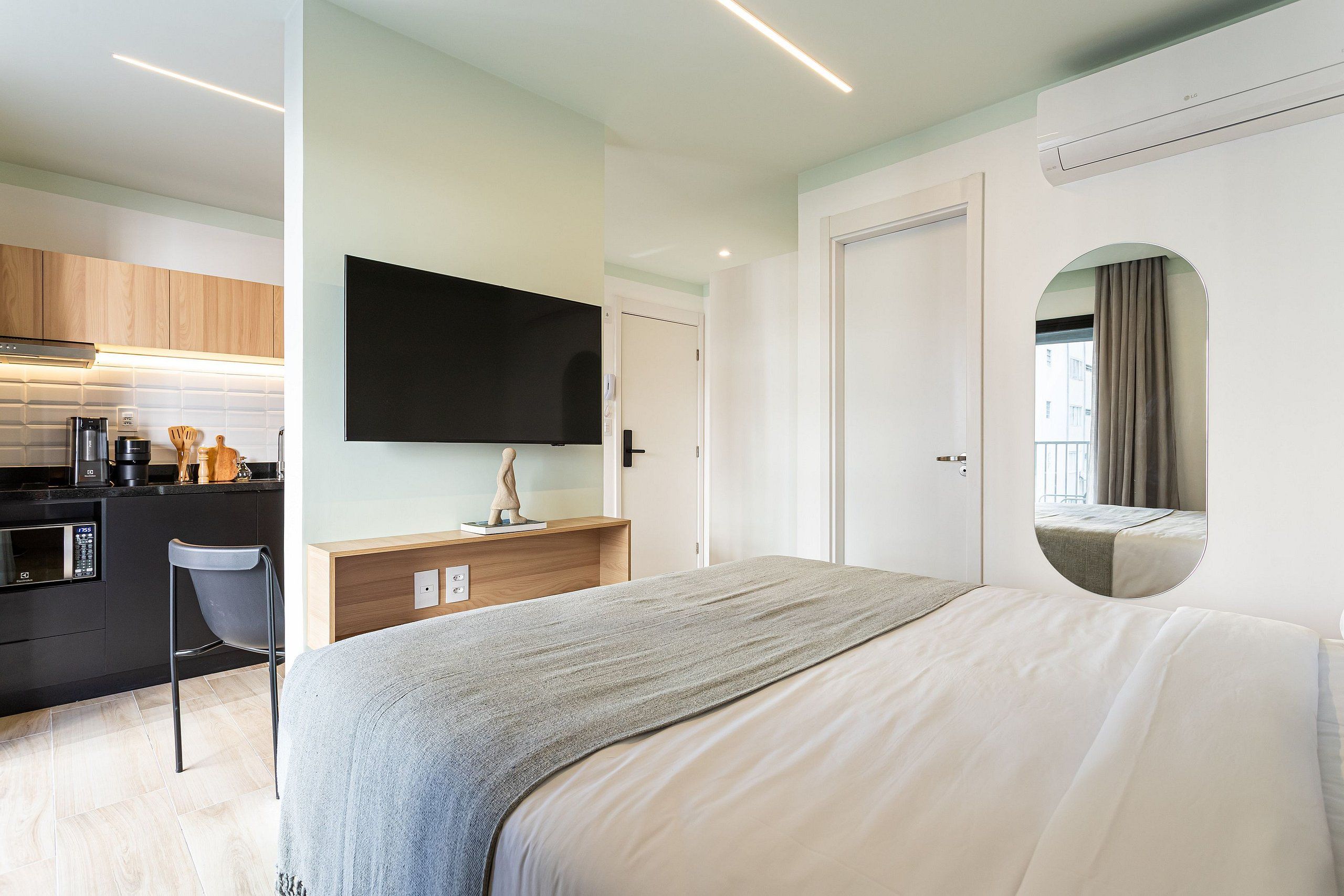 Een moderne hotelkamer met een bed, een flatscreen televisie aan de muur, een klein keukenhoekje en een badkamer met spiegel.