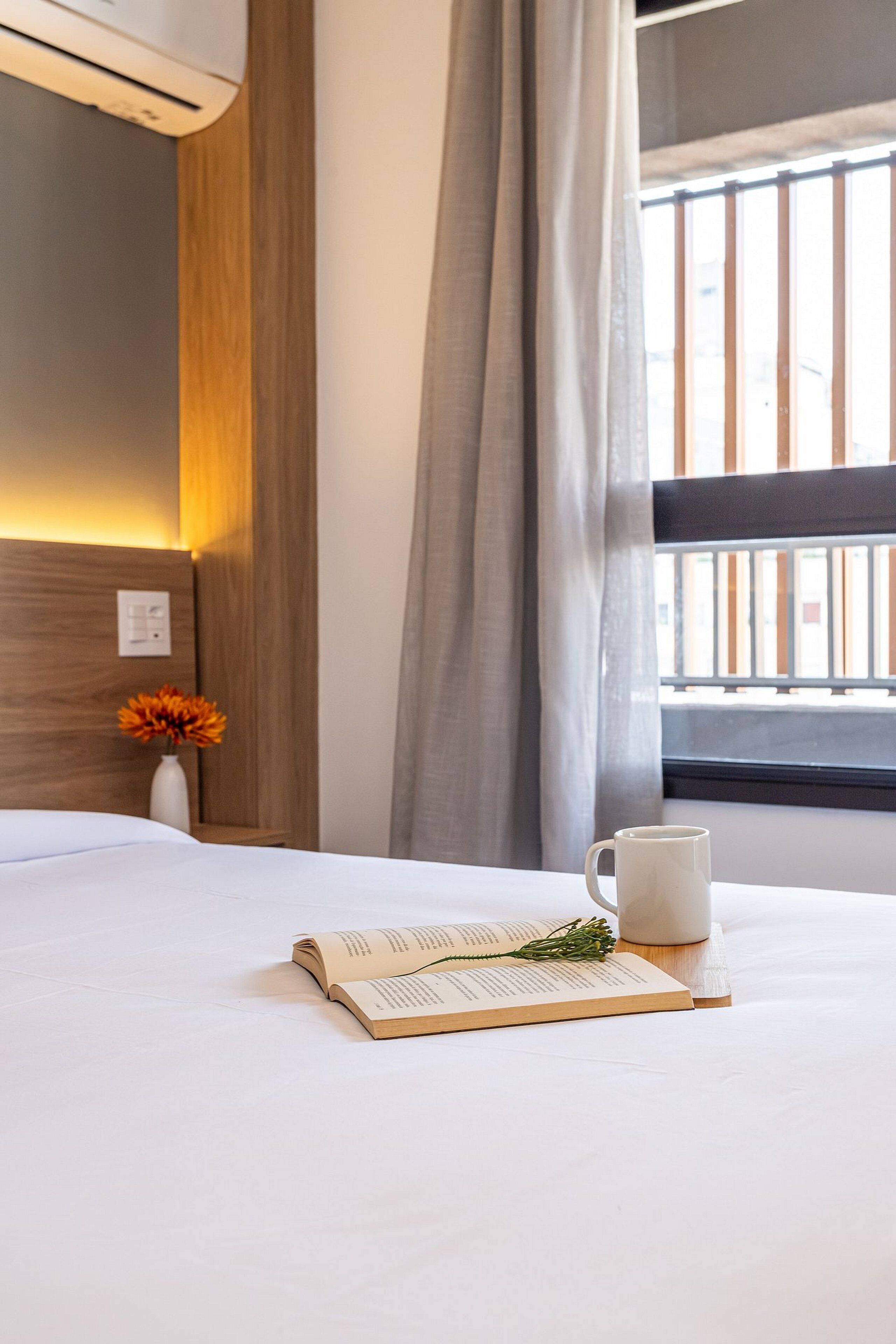 Una cama bien tendida con un libro y una taza, un jarrón con flores, y una ventana con cortinas.
