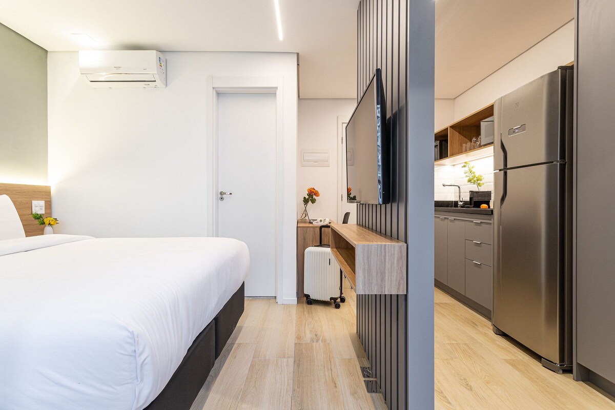 A hotel room with a bed, a desk, a kitchenette with a refrigerator, and an air conditioning unit.