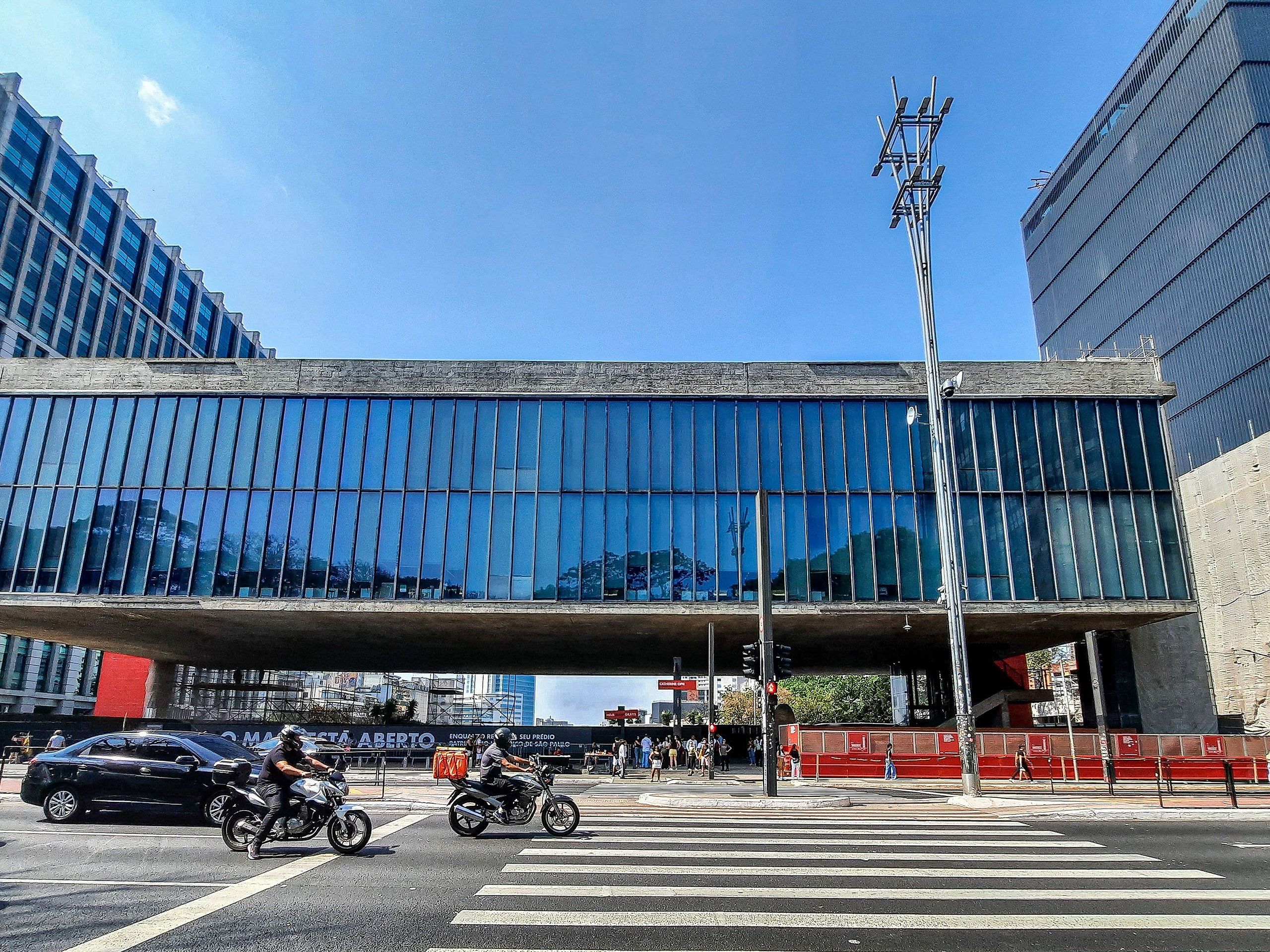 A modern building with a large glass facade, a pedestrian overpass, and a busy street with vehicles and pedestrians.