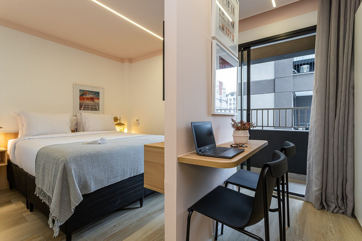 A hotel room with a bed, bedside table, lamp, laptop, and a view of the city through a window.