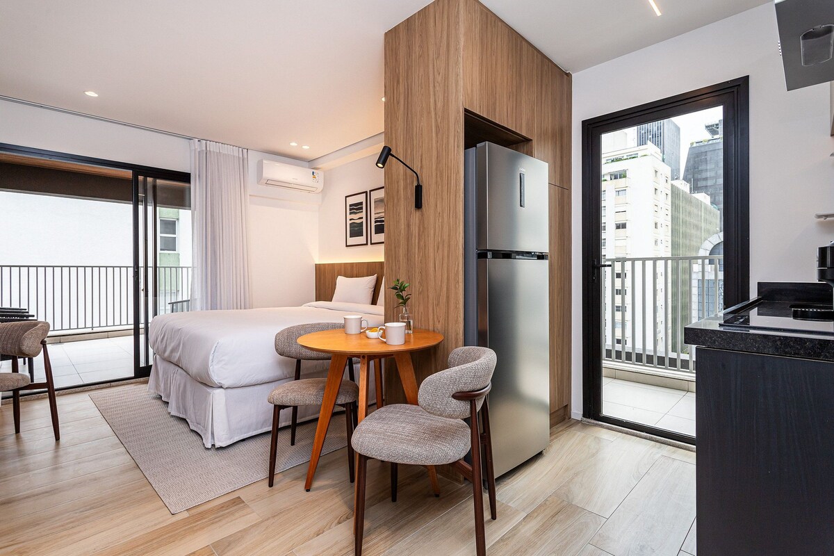A modern hotel room with a bed, a dining table with chairs, a refrigerator, and a view of the city through a large window.