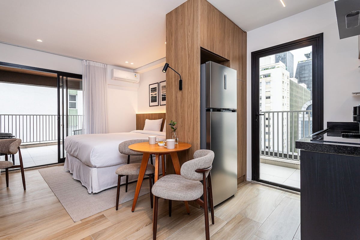 Une chambre d’hôtel moderne équipée d’un lit, d’une table à manger avec des chaises, d’un réfrigérateur et offrant une vue sur la ville grâce à une grande fenêtre.