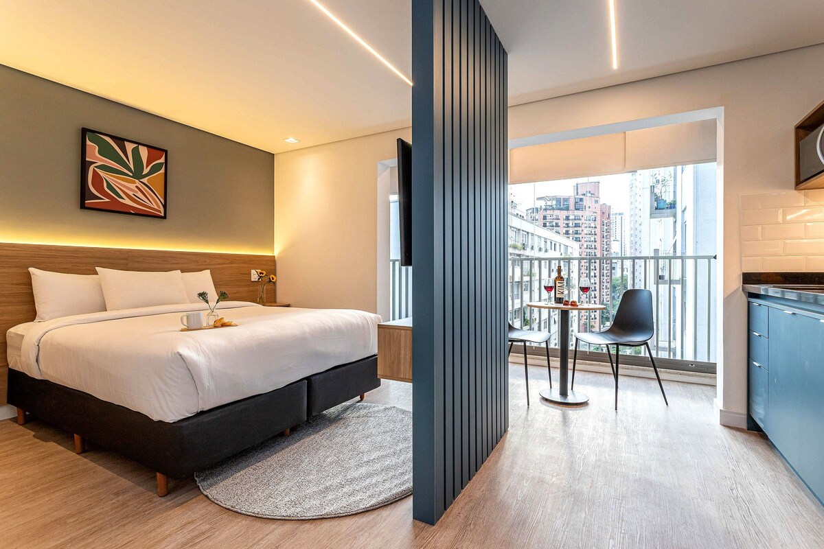 A modern hotel room with a bed, a small table with chairs, a balcony, and a city view.