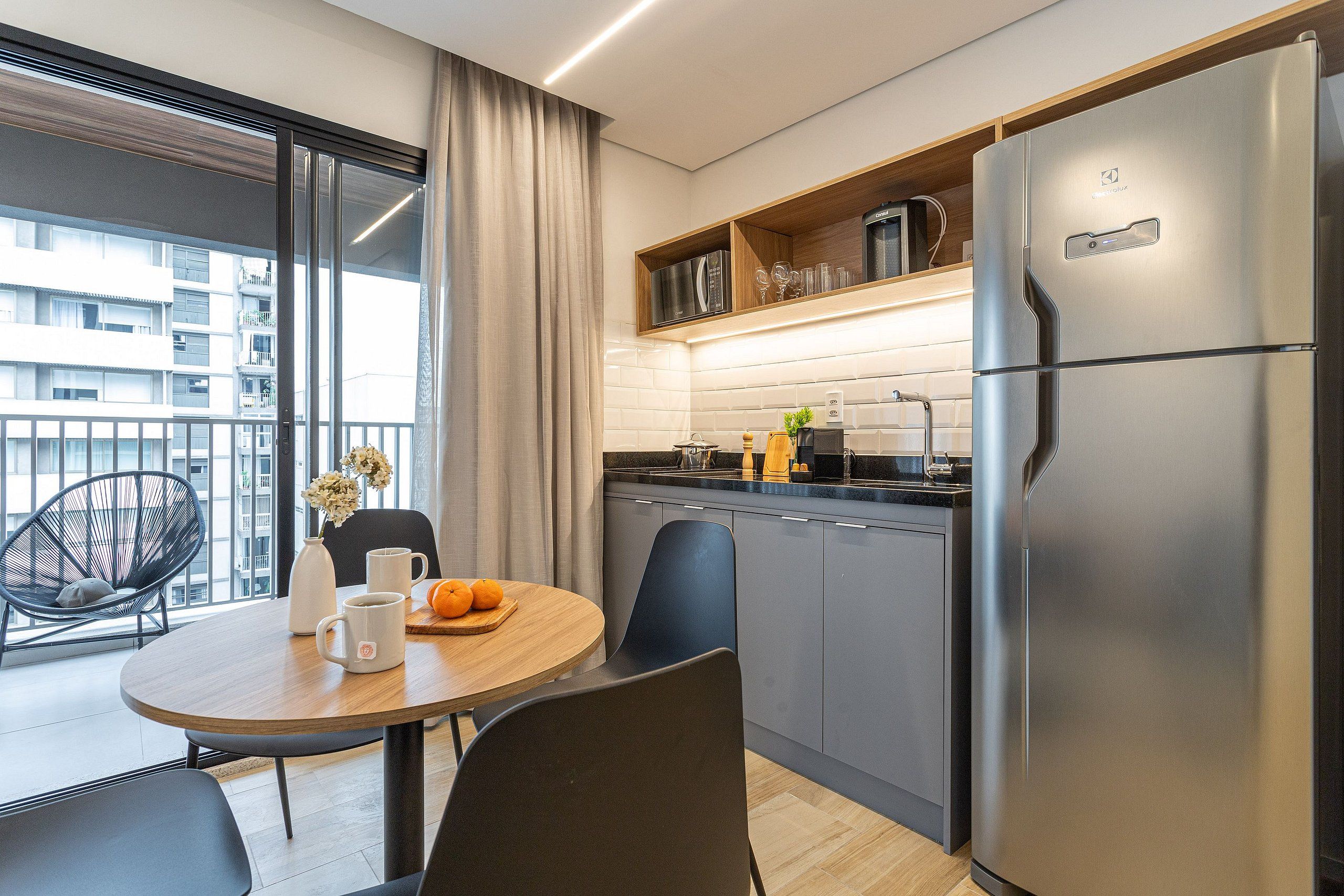 A modern kitchen with a large refrigerator, a dining table with chairs, and a view of the city through large windows.