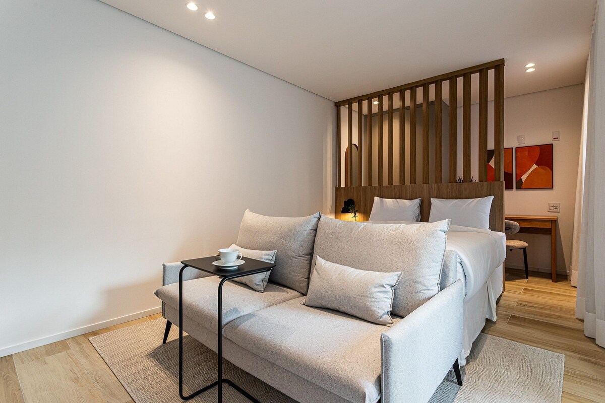 A modern hotel room with a sofa, a small table with a cup, a wooden partition, a desk, and a chair.