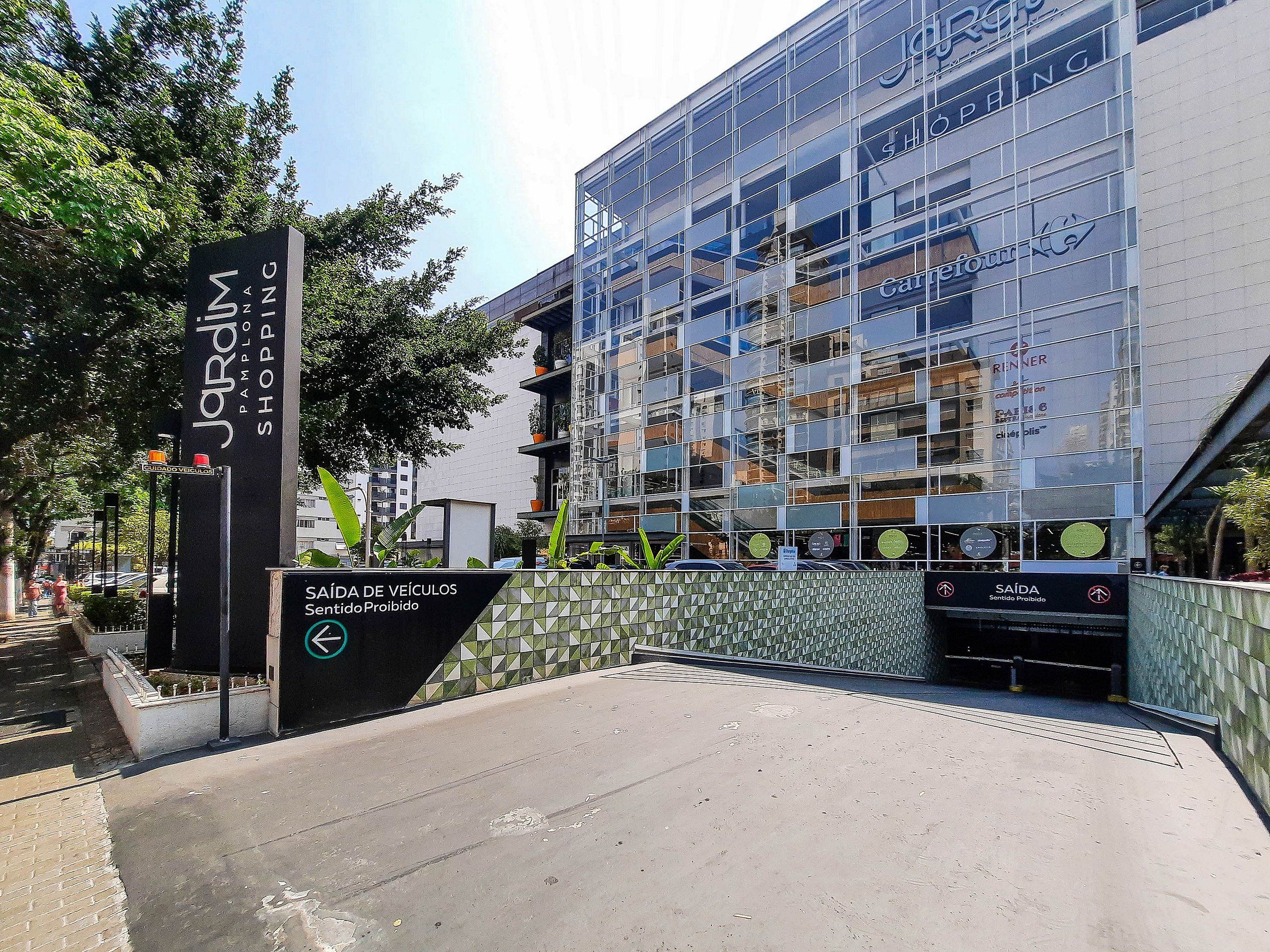 A modern building with a glass facade, a sign for 'Jardim Shopping,' and a 'Saída de Veículos Sentido Proibido' sign.