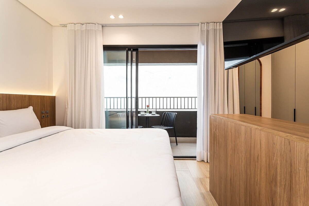 A hotel room with a bed, a wooden desk, a balcony with a table and chairs, and a sliding door.
