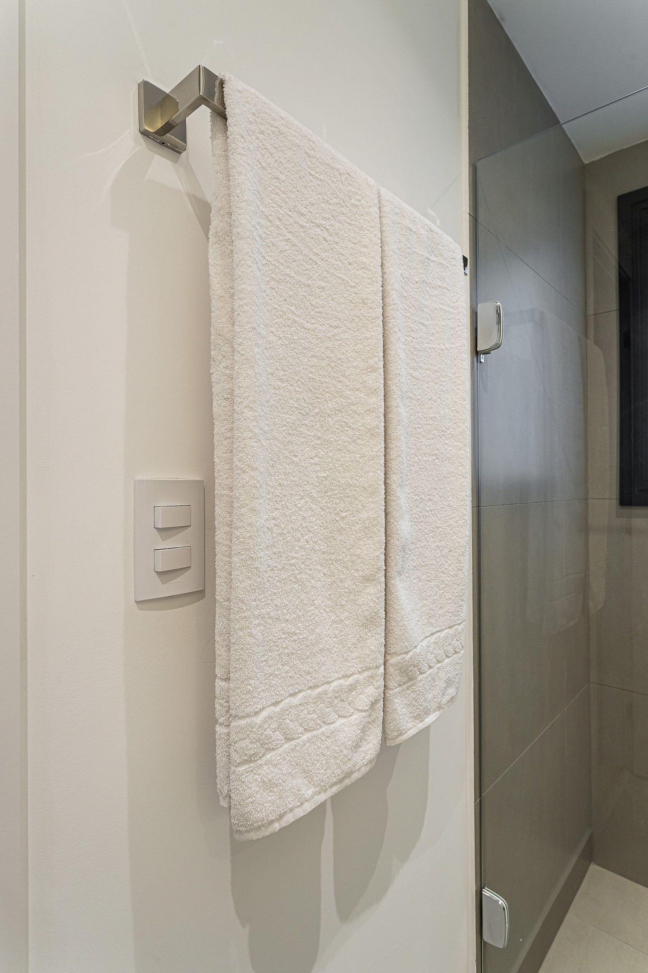 A bathroom with a white towel hanging on a metallic towel rack, a glass shower door, and a light switch on the wall.