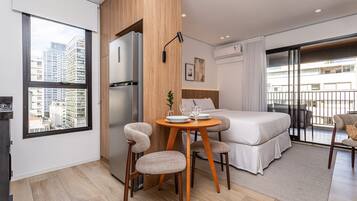 Ett modernt hotellrum med en säng, ett matbord med stolar, ett kylskåp och utsikt över stadsbilden.
