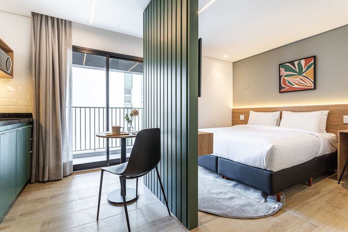A hotel room with a bed, a chair, a small table, a window with curtains, and a balcony.