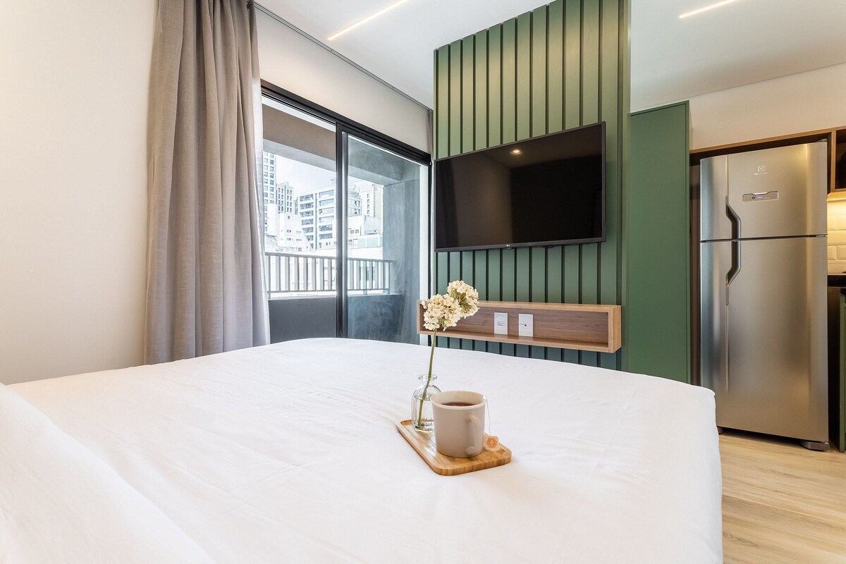 A hotel room with a bed, a TV mounted on the wall, a refrigerator, and a window with curtains.