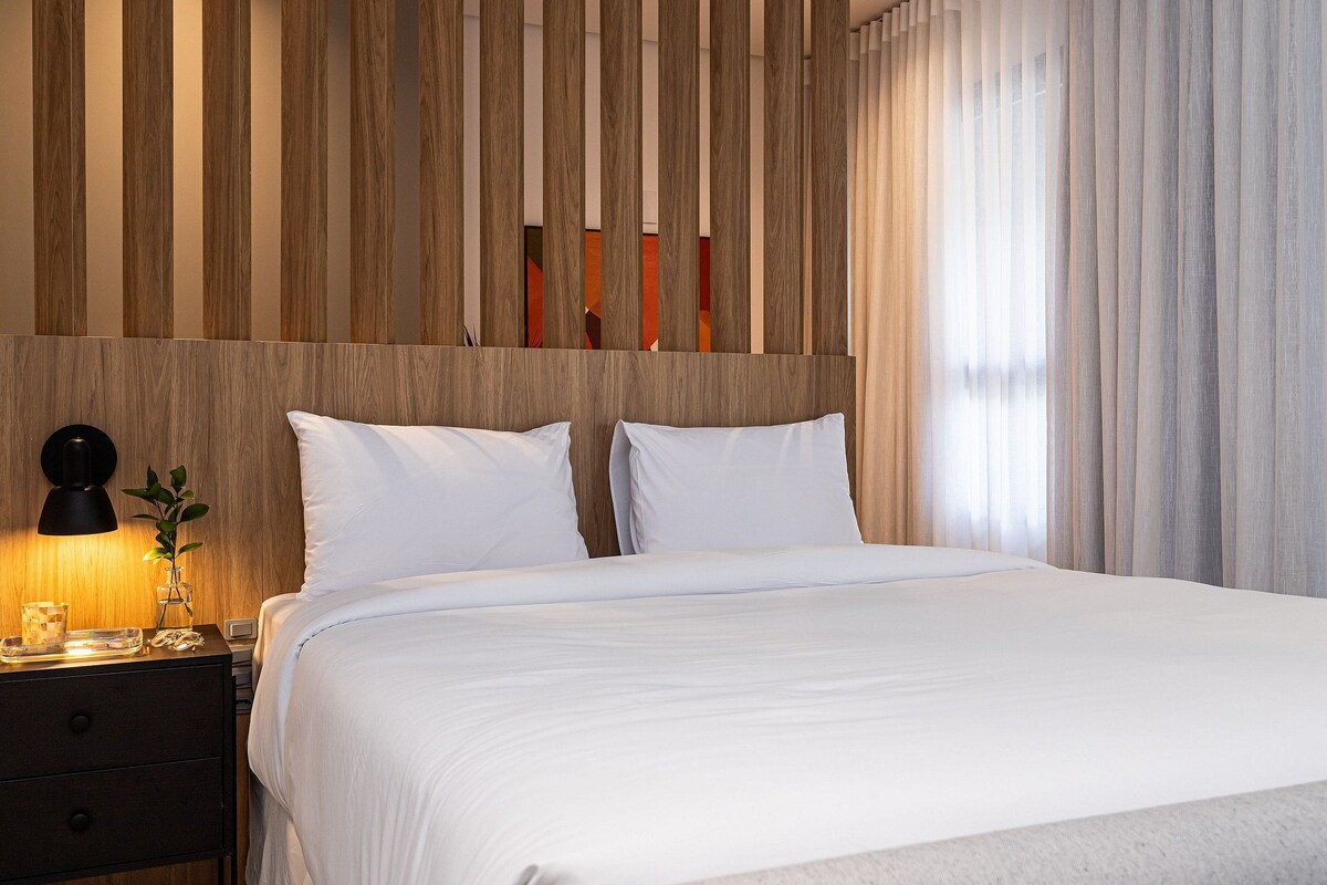 A hotel room with a wooden headboard, a bed with white linens, a bedside table with a lamp, and a window with sheer curtains.