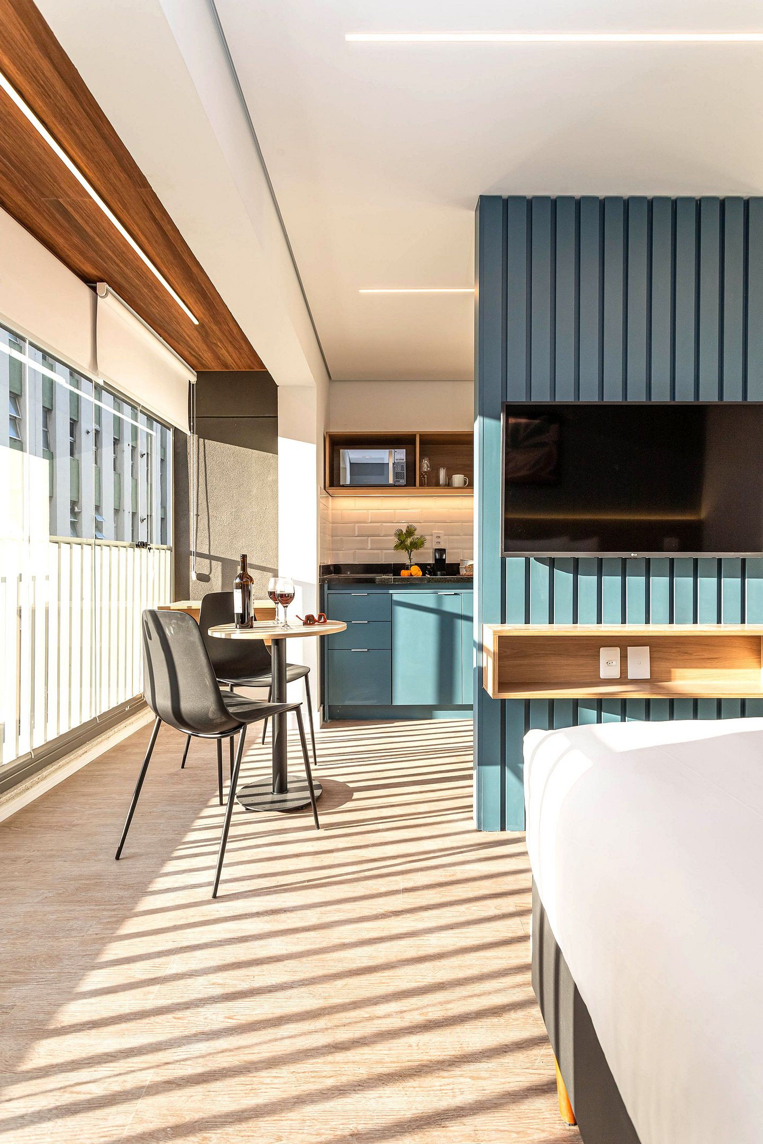 Una moderna camera d'hotel con un letto, un angolo ristoro con tavolo e sedie, una cucina e un grande televisore montato a parete.