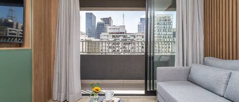 A modern living room with a grey sofa, a small round table with a vase of flowers, and a view of a cityscape through large windows.