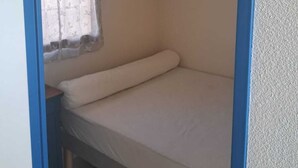 A small, single-bed room with a window covered by a sheer curtain, a bedside table, and a hanging light fixture. - 1-bedroom studio with sea view. Ideal for 2 adults and 1 child. (Saint Pierre la mer)