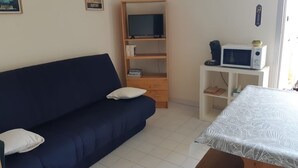 A room with a blue sofa, a wooden TV stand, a microwave, and a bed with a patterned bedspread. - 1-bedroom studio with sea view. Ideal for 2 adults and 1 child. (Saint Pierre la mer)