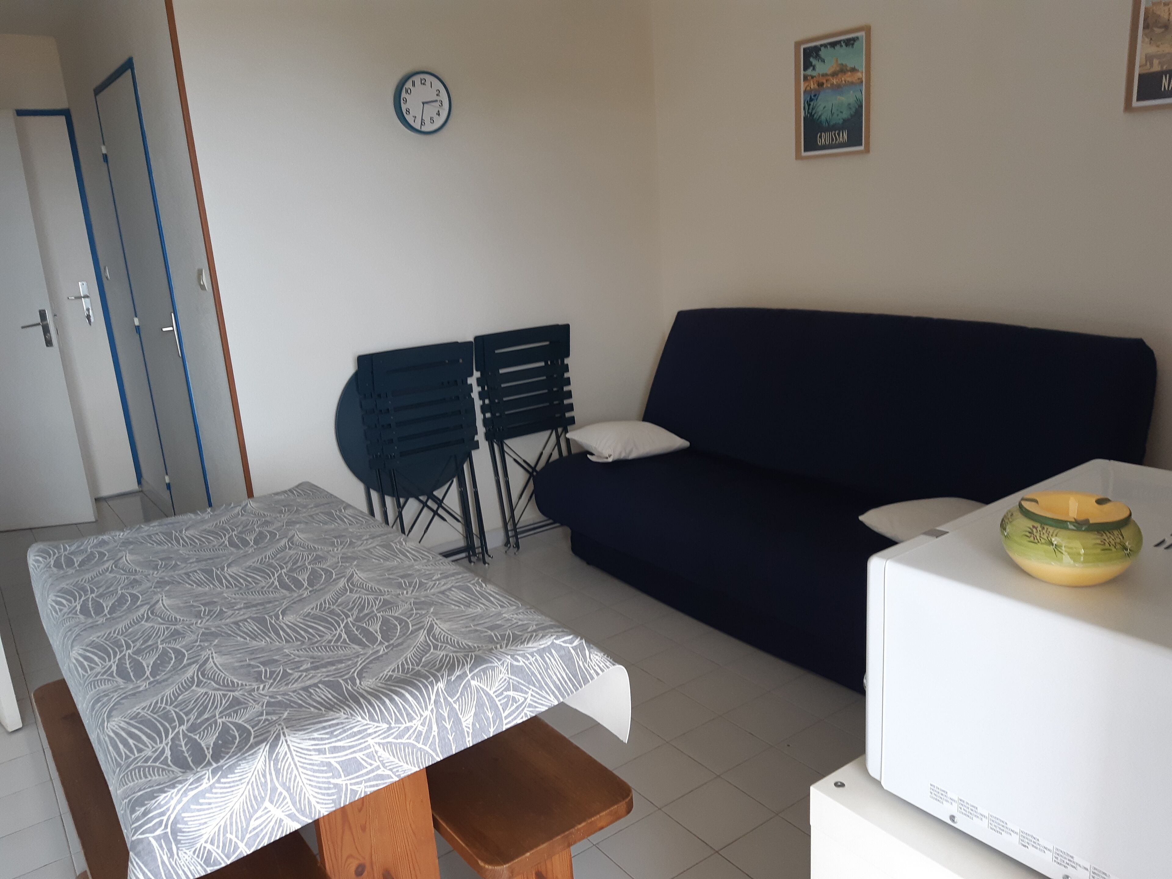 Une pièce comprenant un lit, un canapé bleu foncé, une petite table avec un bol, et une horloge au mur.
