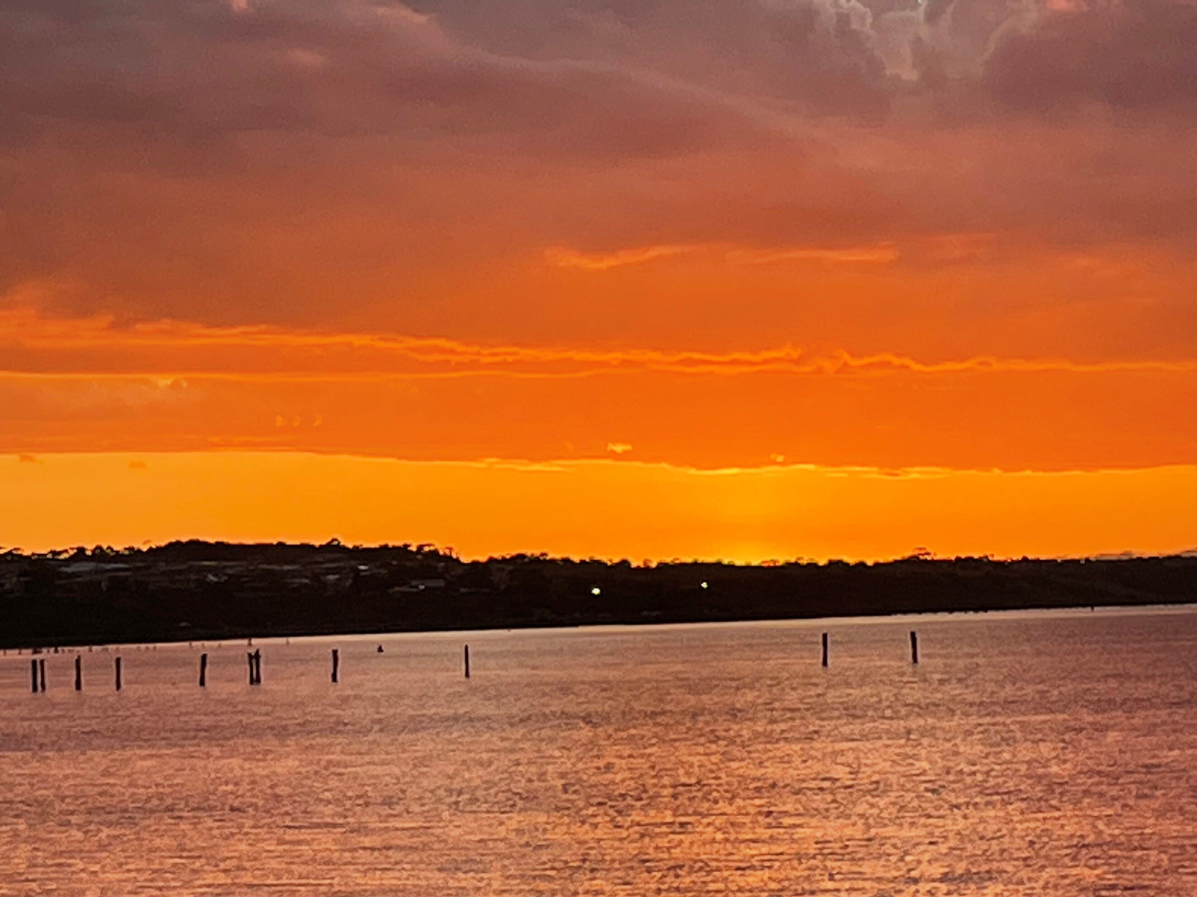Un atardecer sobre una superficie acuática tranquila, con una costa lejana y varias estacas de madera.