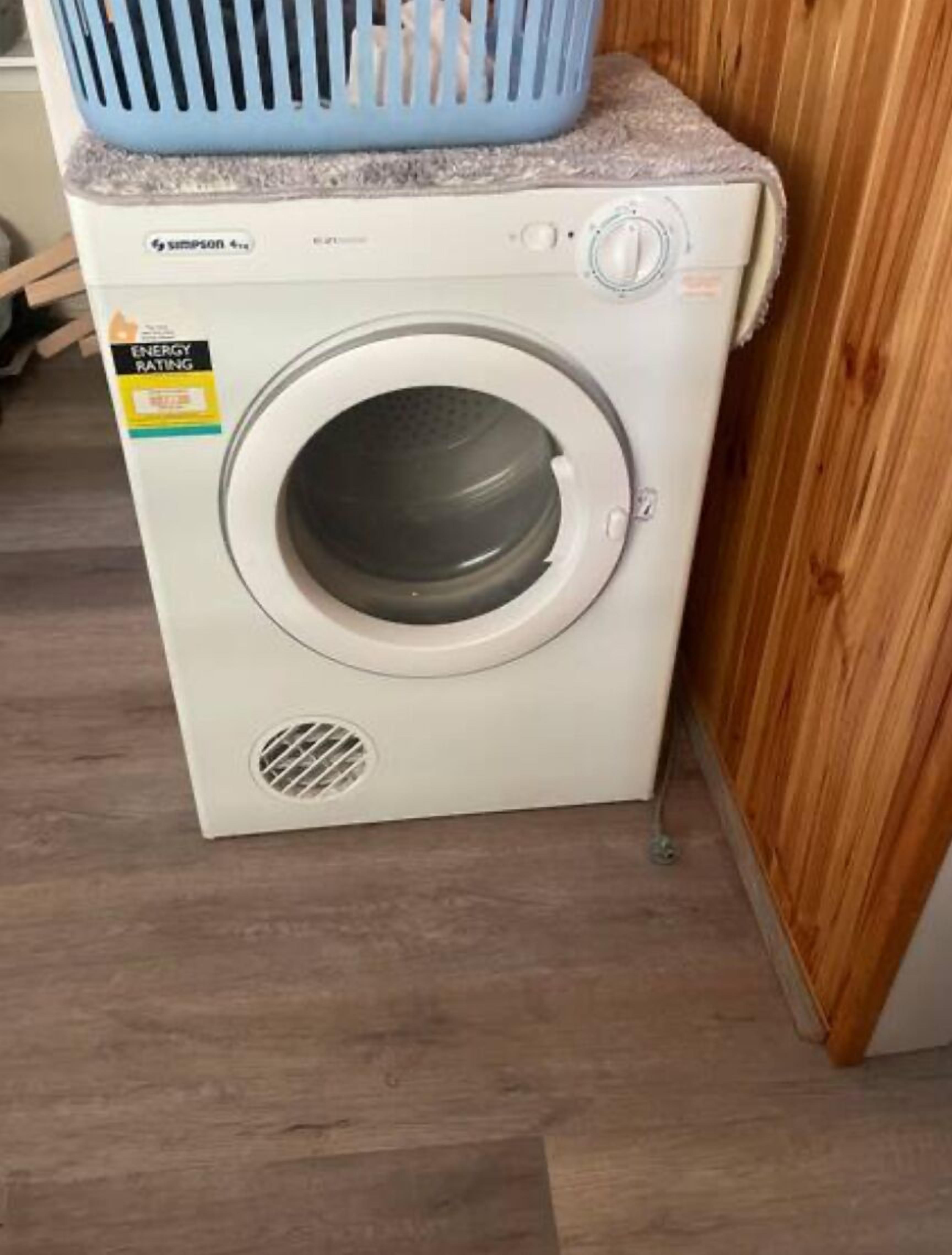 A front-loading washing machine with a blue basket on top, placed in a corner with wooden flooring.