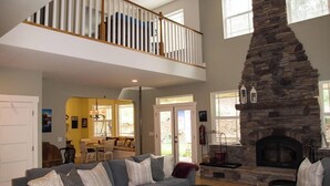 A spacious living room with a stone fireplace, a high ceiling with a fan, and a dining area in the background.