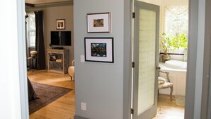 A hallway with a wooden floor, a door leading to a bathroom, and a television on a stand in the adjacent room.