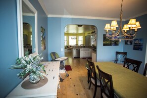 A dining area with a table, chairs, and a chandelier. There is a kitchen visible in the background.
