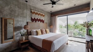 Une chambre moderne avec un grand lit, un ventilateur de plafond, une vue sur la verdure et une table de chevet en bois surmontée d’un vase.