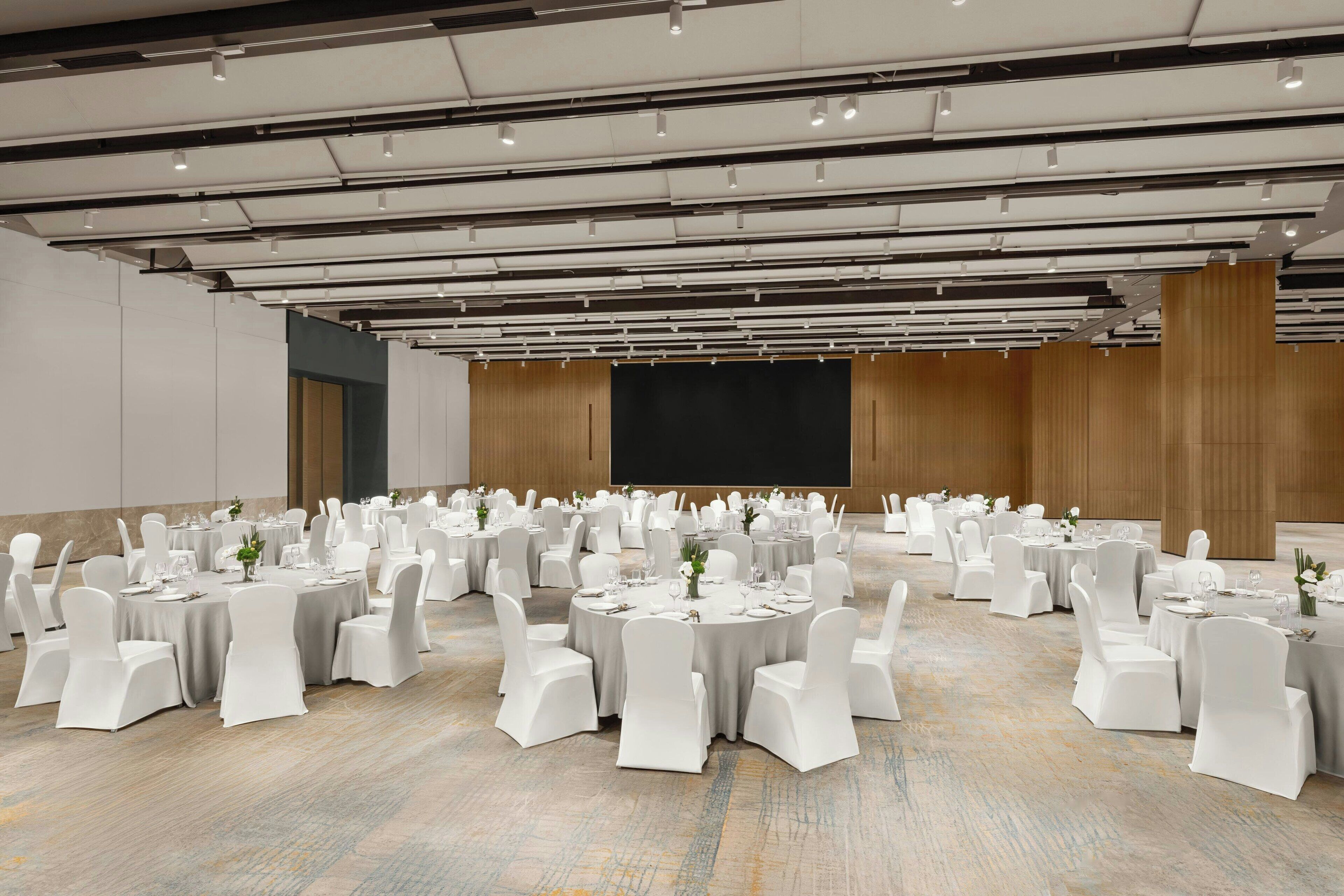 A large conference room with round tables and white chairs, equipped with overhead lighting and a large screen.