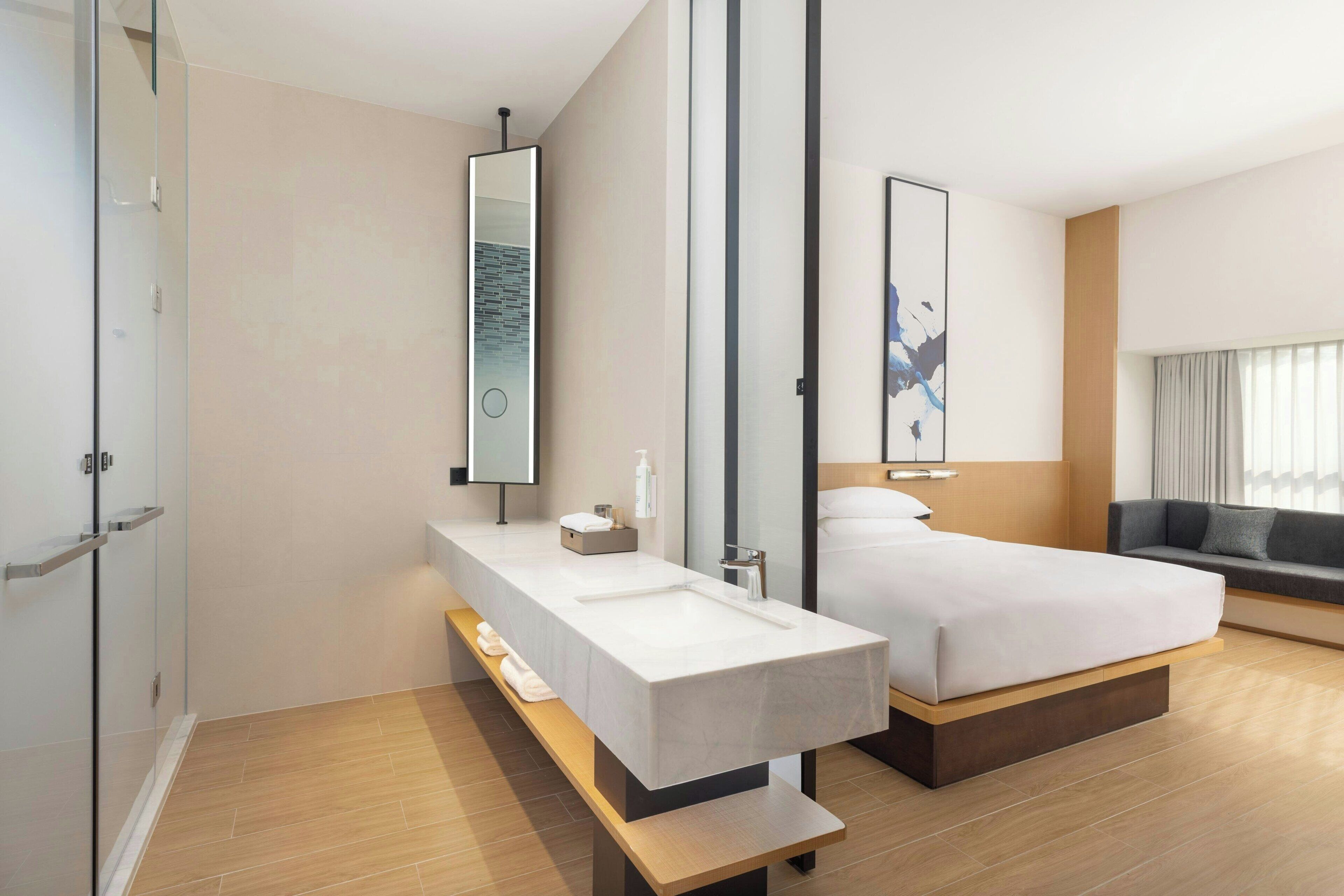 Quarto de hotel moderno com uma bancada de banheiro espaçosa, uma cama, um sofá e um espelho grande.