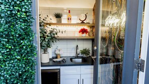 Une cuisine avec des meubles blancs, un plan de travail noir et divers éléments décoratifs, notamment des plantes et un panneau lumineux néon.