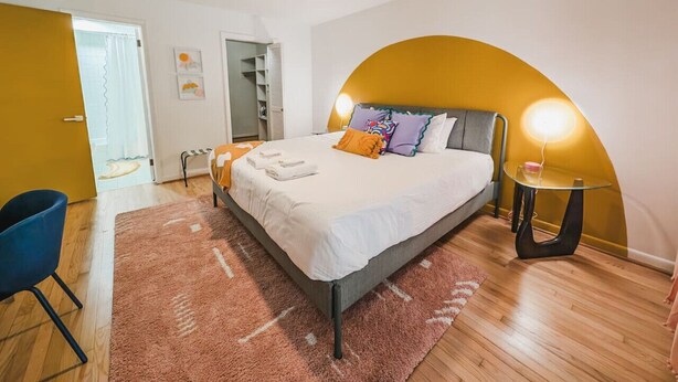 A modern bedroom with a bed, a blue chair, a glass table, and a unique curved wall design.