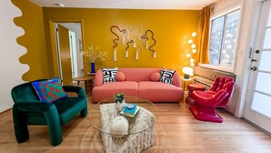 A living room with a yellow accent wall, a green sofa, a pink sofa, a glass coffee table, and a red chair.