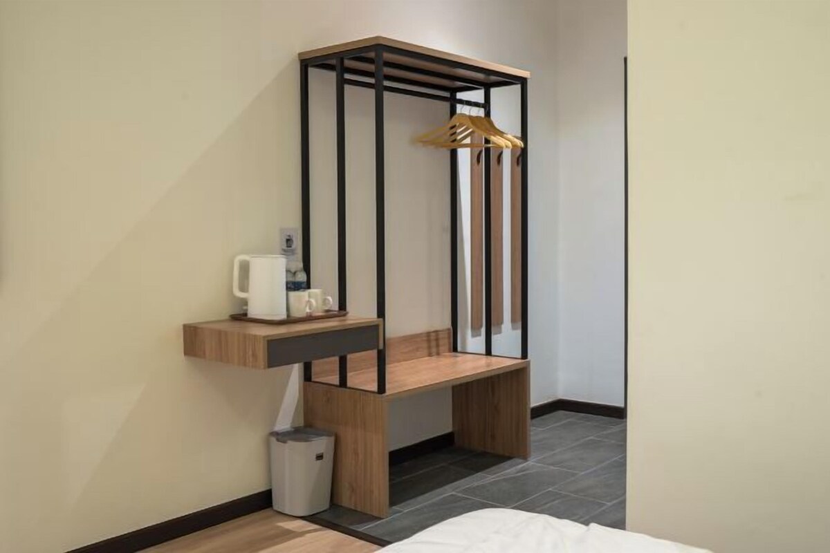 Habitación pequeña con una cama, un estante de madera con una tetera, un cubo de basura y un perchero.