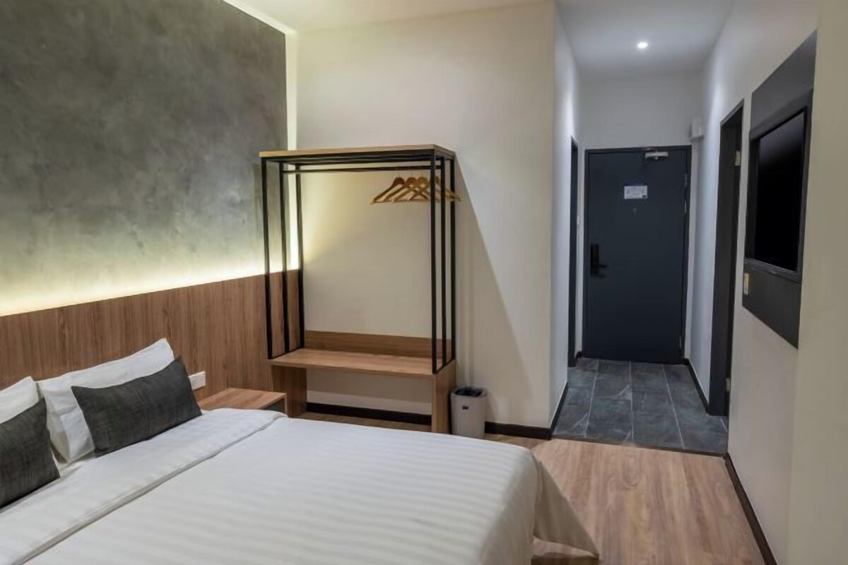 A hotel room with a bed, a wooden headboard, a wall-mounted TV, and a hallway leading to a door.
