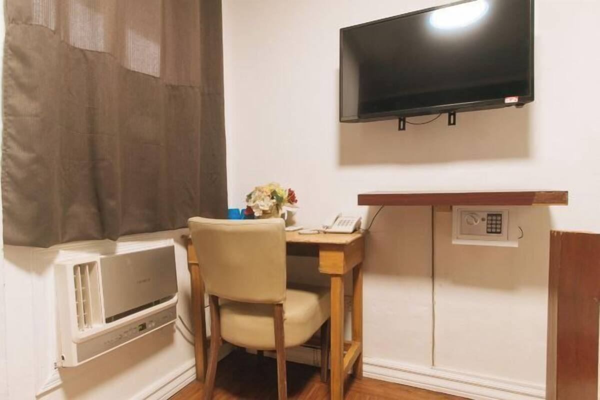 Una habitación pequeña con una cama, una mesita de noche, un televisor en la pared y una ventana con cortinas.
