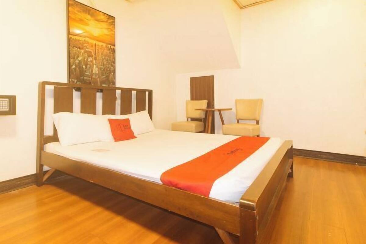 Una cama bien hecha con estructura de madera, colchón blanco y manta roja. Junto a la cama, una mesita redonda con una silla.
