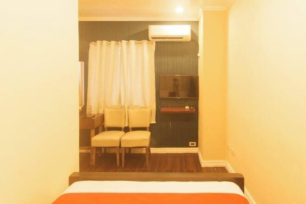 Habitación de hotel con cama, dos sillas, una mesita pequeña, equipo de aire acondicionado y televisor.