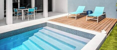 Un área de piscina moderna al aire libre con un deck de madera, dos sillones de descanso y un juego de comedor.
