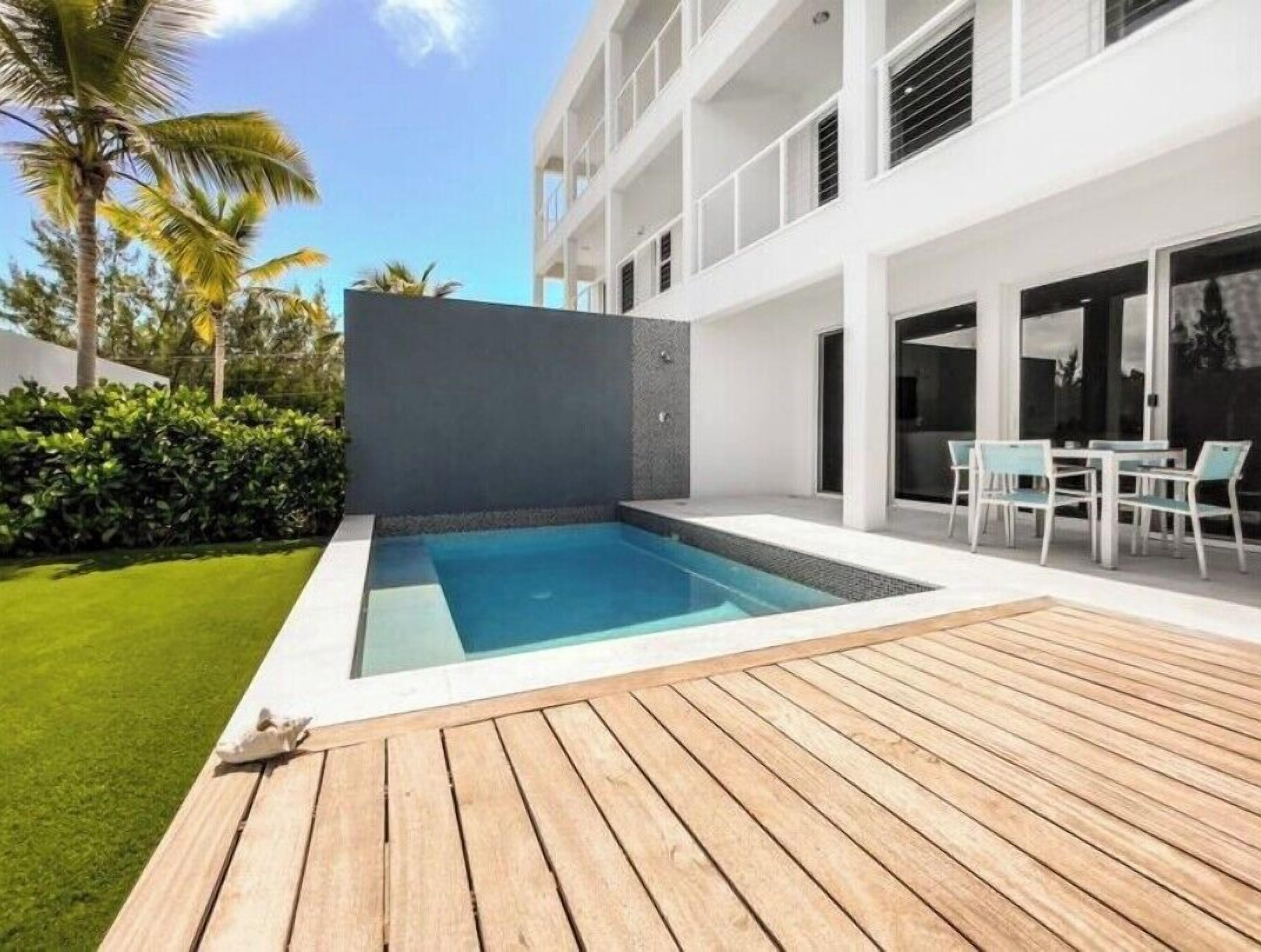 Un área de piscina moderna al aire libre con un deck de madera, rodeada por un c ésped cuidado y palmeras.