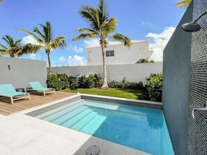 Una piscina moderna al aire libre con sillones de descanso, palmeras y un edificio blanco al fondo.
