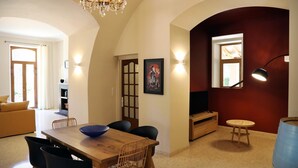 A dining area with a wooden table, chairs, and a chandelier. There is a television on a stand and a small round table with a lamp. - Gîtes de France® - Gîte du Chateau Saint Didier (PARNAC)