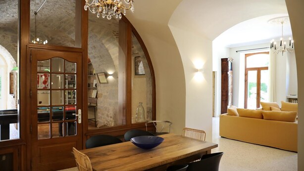 A dining area with a wooden table, chairs, and a chandelier. There is a sofa in the background and a doorway leading to another room. - Gîtes de France® - Gîte du Chateau Saint Didier (PARNAC)