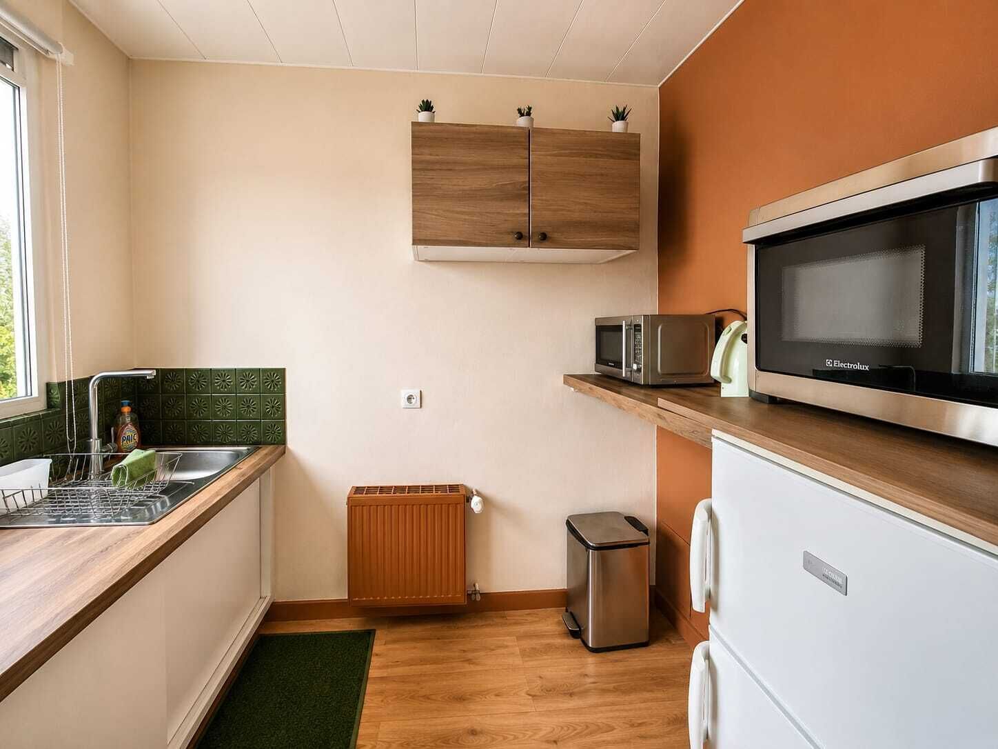 A compact kitchen with a microwave, a small trash bin, a radiator, and a wooden cabinet above the sink.