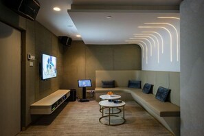 Un área de descanso moderna con un sofá curvo, un televisor de pantalla plana, un quiosco de información digital y una zona de estar con mesas redondas.
