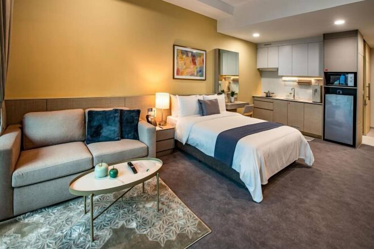 A hotel room with a bed, sofa, nightstand, lamp, and kitchenette.