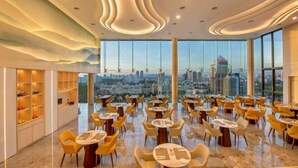 A modern dining area with round tables and yellow chairs, large windows offering a city view, and a unique ceiling design. - Sunway Sanctuary - Seniors Hotel & Residences (Subang Jaya)