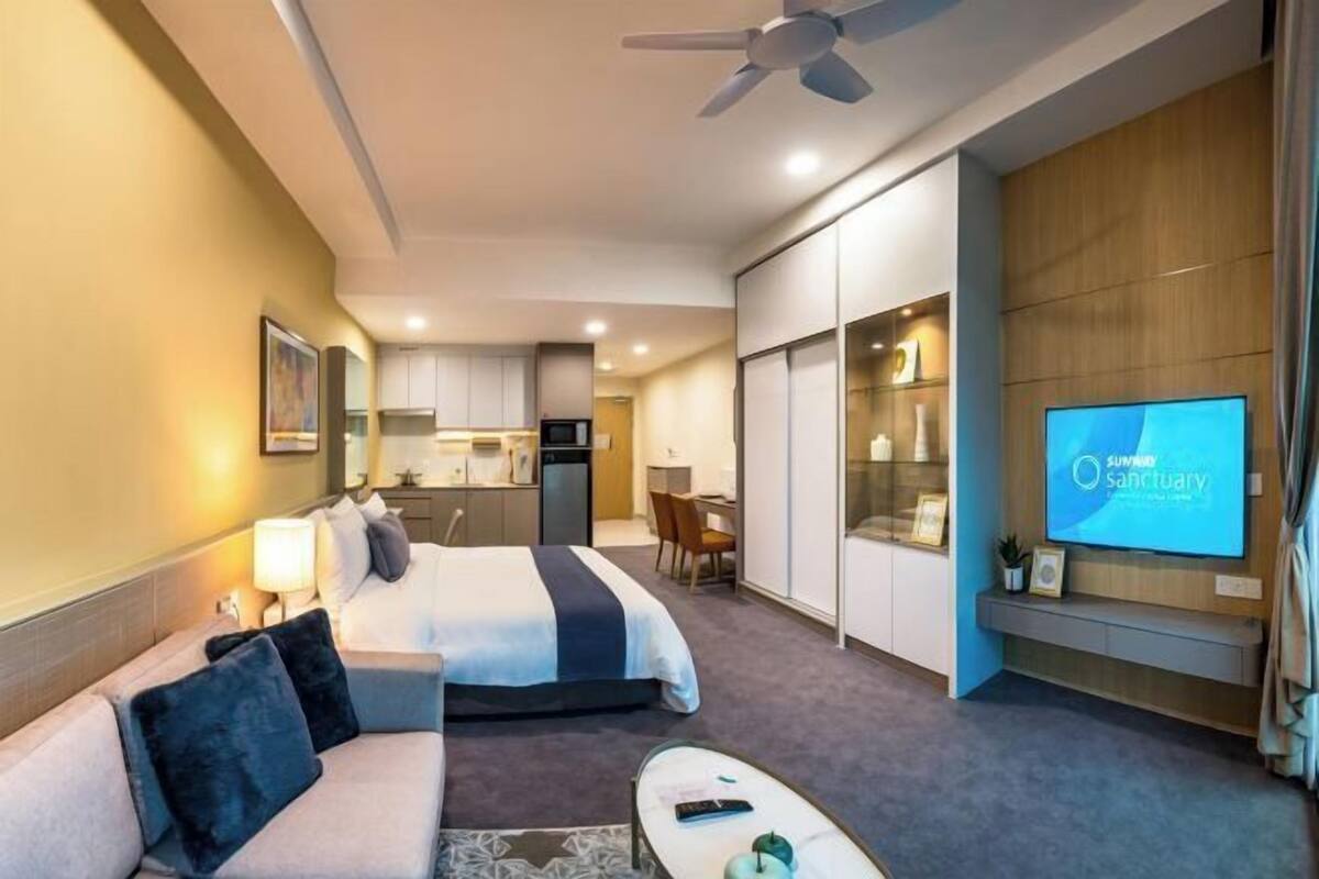 A hotel room with a bed, a sofa, a desk, a television, and a kitchenette in the background.