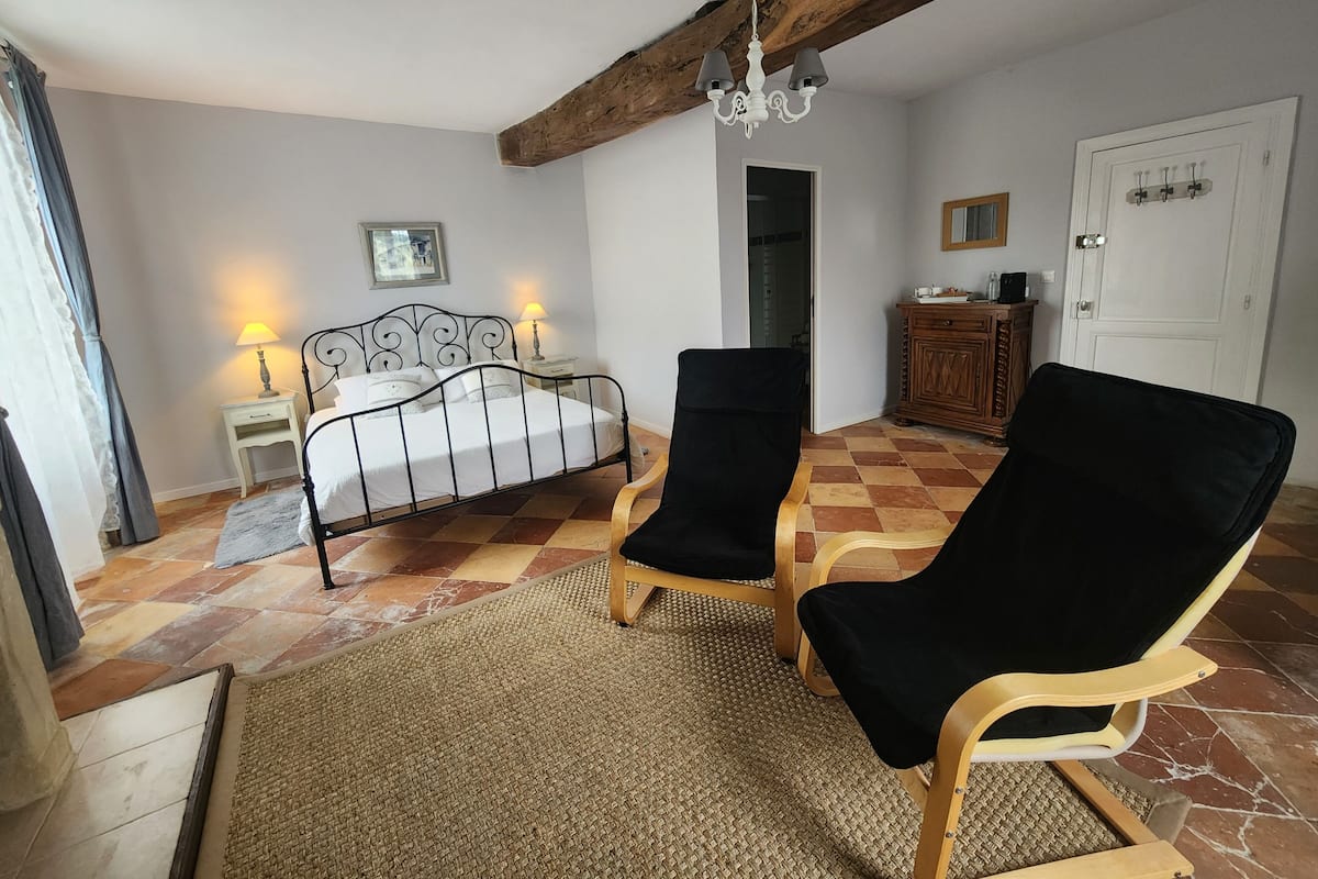 Un dormitorio con cama, dos sillas, una mesita de noche, una lámpara, un armario de madera y una puerta.